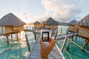 a hand holding a phone to a dock with huts on the water