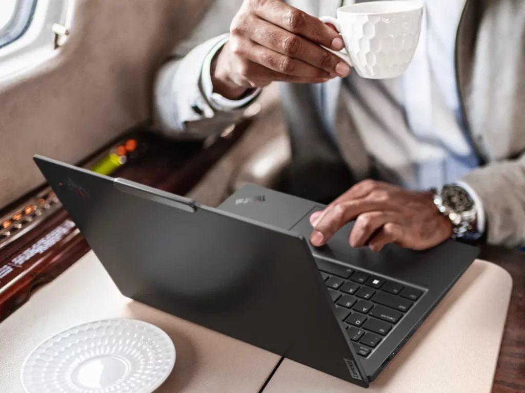 a person holding a cup of coffee and typing on a laptop. Lenovo ThinkPad X1 Carbon Gen 12.  {Tech} for Travel. https://techfortravel.co.uk