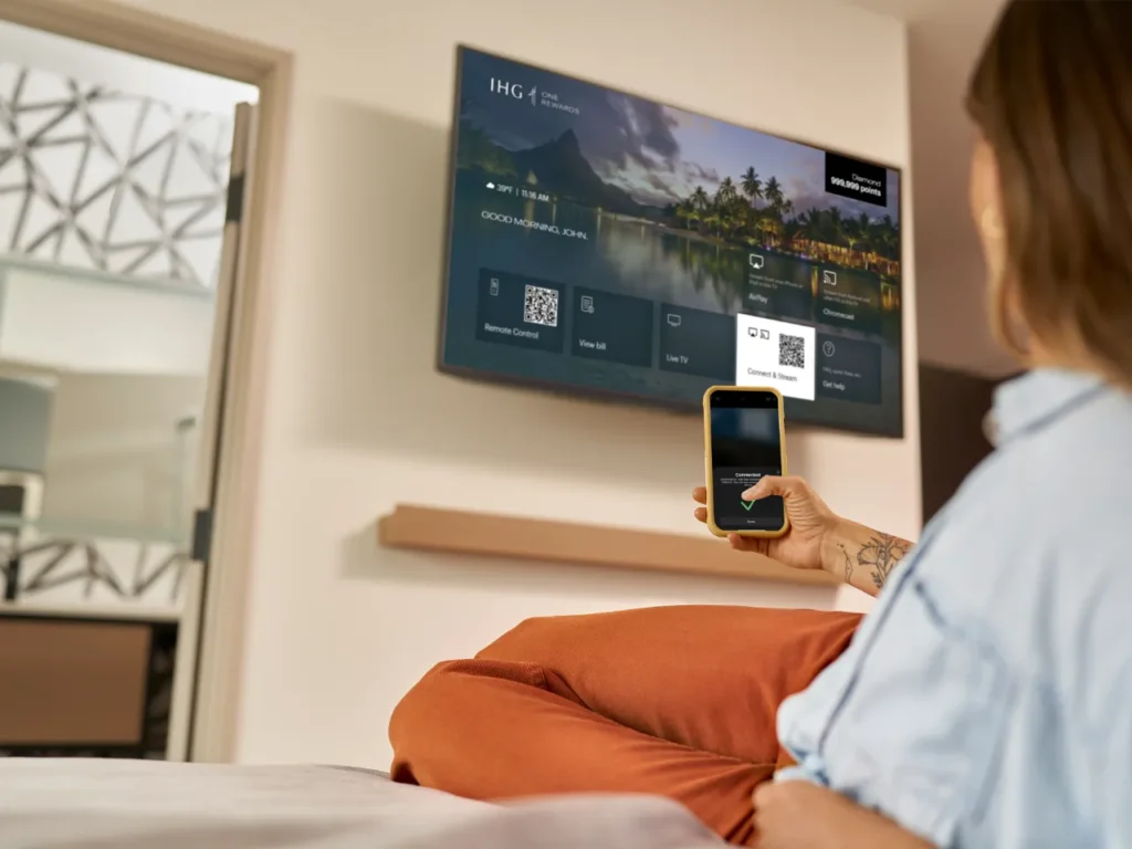 a woman sitting on a bed looking at a television screen. IHG AirPlay hotels. {Tech} for Travel. https://techfortravel.co.uk