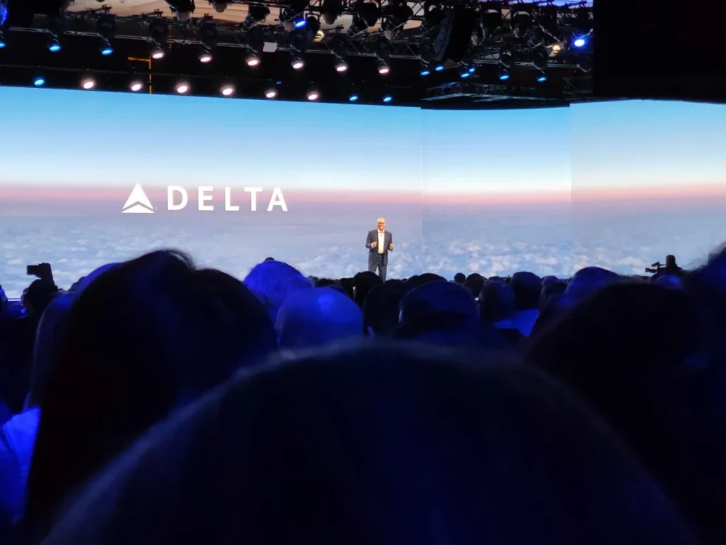 a man standing on a stage with a large screen with a large crowd. Delta CES Keynote.  {Tech} for Travel. https://techfortravel.co.uk