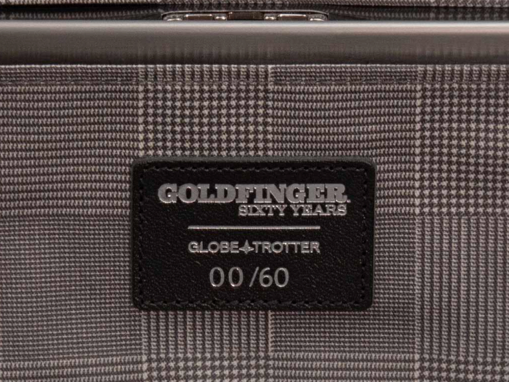 a black leather label on a grey and white checkered bag. Globe-Trotter 007 Goldfinger Limited Edition Carry On. {Tech} for Travel. https://techfortravel.co.uk