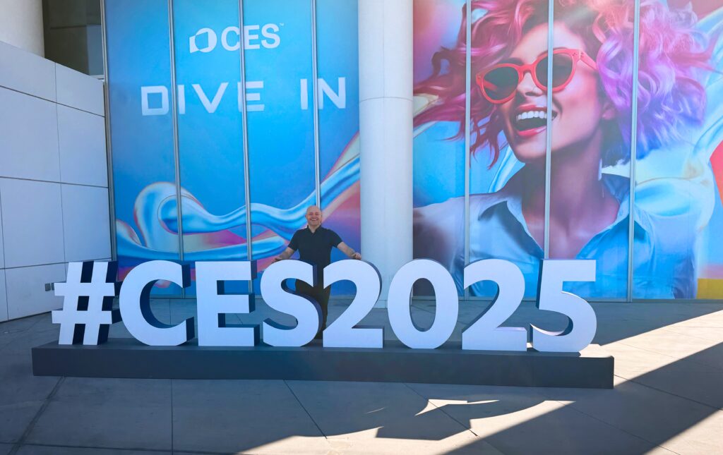 a man standing in front of a large sign. Best Travel Gadgets 2025. Best of CES 2025. {Tech} for Travel. https://techfortravel.co.uk