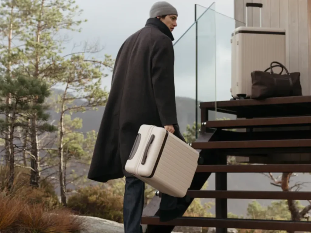 a man in a long coat carrying a suitcase. Carl Friedrik Carry-On Core. Best Carry On Cases 2025. {Tech} for Travel. https://techfortravel.co.uk