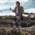 a woman standing on a rocky beach holding a camera. Ascentiz modular exoskeleton. CES 2026 News. {Tech} for Travel. https://techfortravel.co.uk